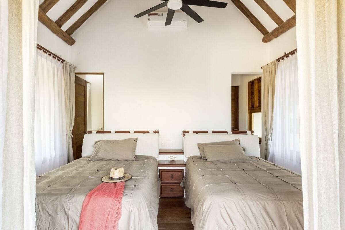 A room with two beds, a ceiling fan, a mirror, and a window with curtains.