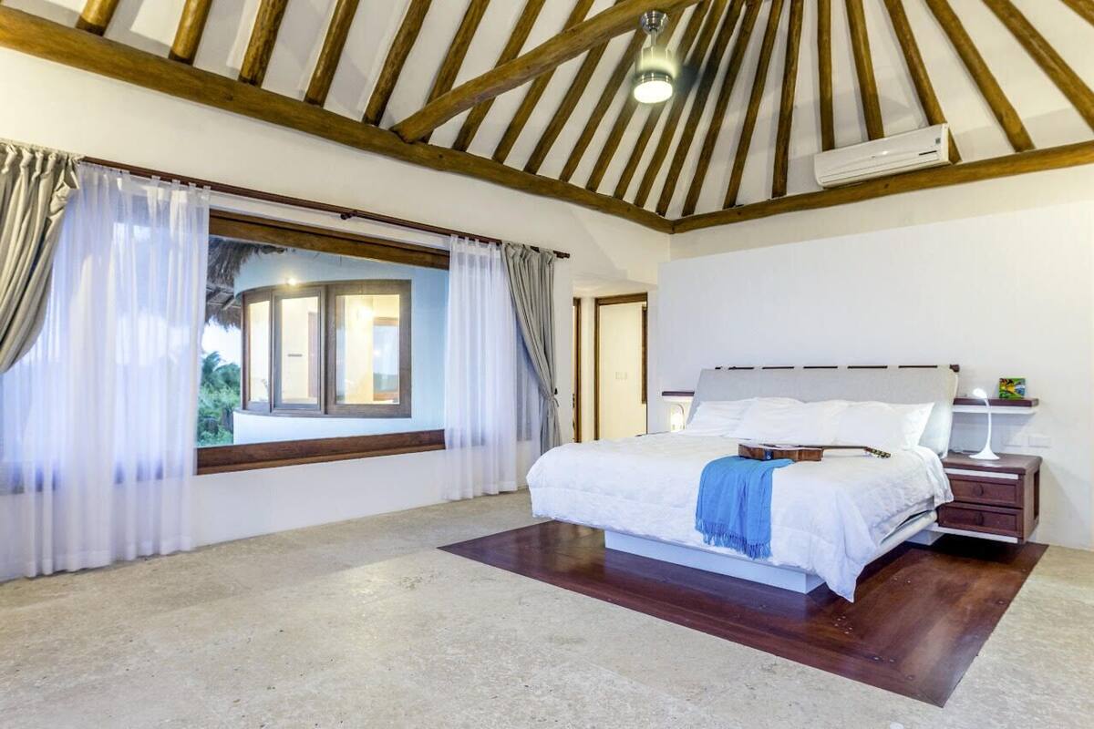 A bedroom with a large bed, a window with curtains, a bedside table with a lamp, and a ceiling fan.
