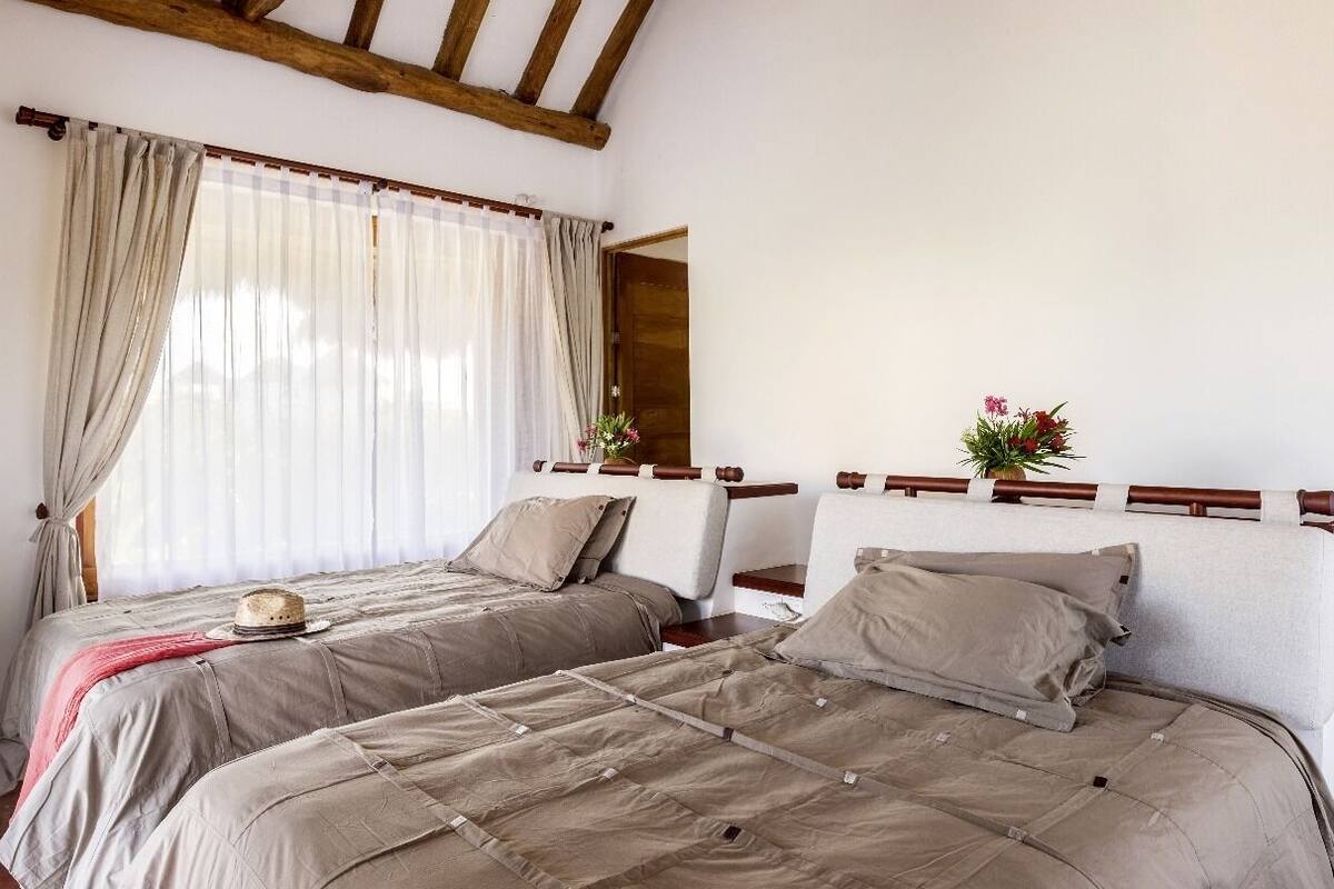 A bedroom with two beds, beige bedding, a wooden headboard, a window with sheer curtains, and a small table with a vase of flowers.