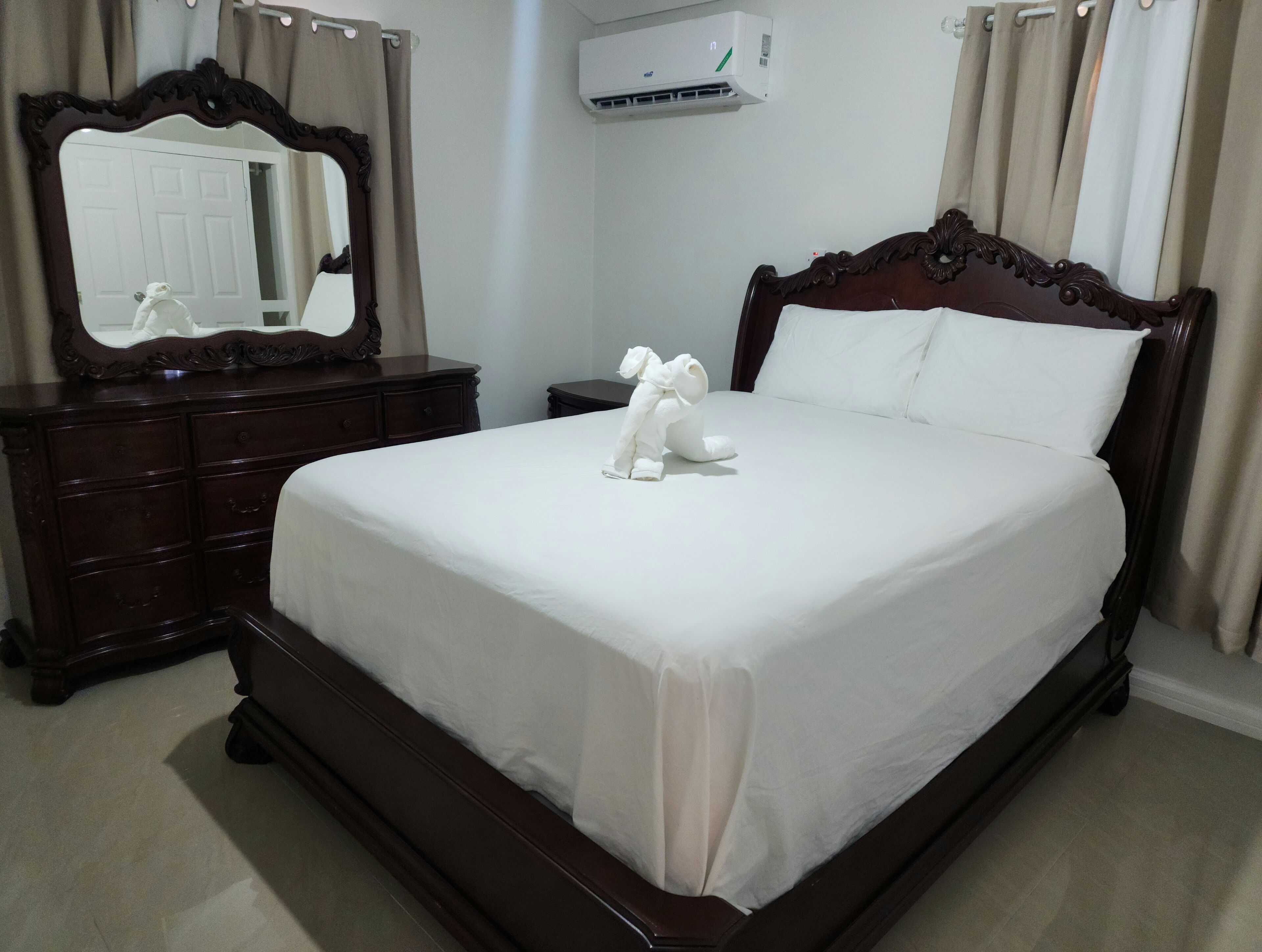 A neatly made bed with a white comforter and pillows, a wooden dresser with a mirror, and an air conditioning unit on the wall.