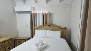 A neatly made bed with white linens and a folded towel, a wooden nightstand with drawers, a wall-mounted air conditioner, and a curtain. - PLAN YOUR GETAWAY UNDER THE SUN 
           Dazzle Key Suite (Saint Ann's Bay)