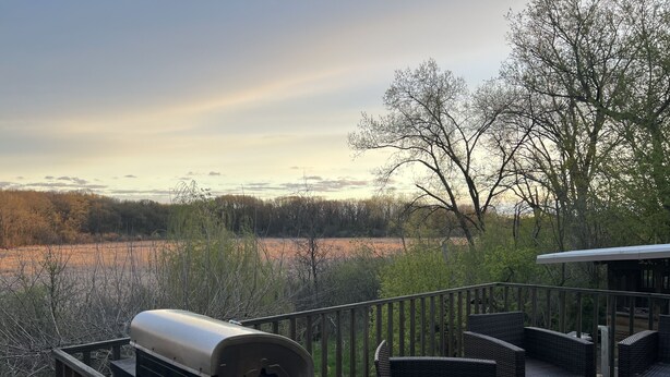 A deck with a grill, wicker furniture, and a view of a field and trees.