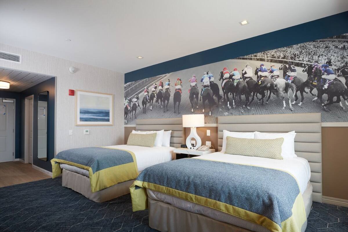 A hotel room with two beds, a large mural of a horse race, a nightstand, and a view of the ocean.
