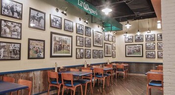 Interior de un restaurante con mesas y sillas de madera, una pared decorada con fotografías blanco y negro, y un letrero verde en la parte superior.