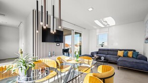 A modern living room with a glass dining table, yellow chairs, a dark sofa with colorful cushions, and a large TV mounted on the wall.