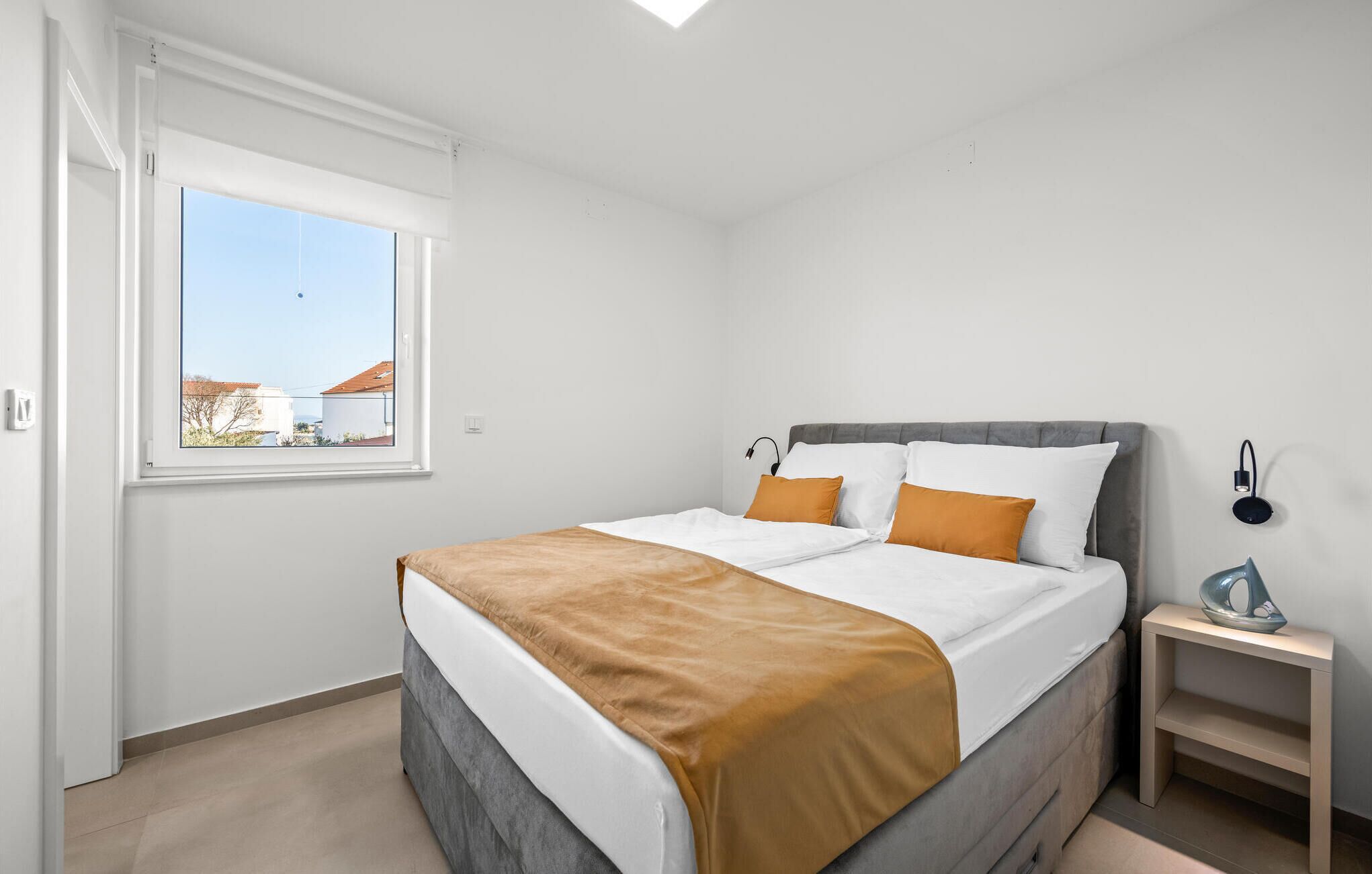 A bedroom with a bed, a bedside table, a lamp, and a window with a view of buildings.