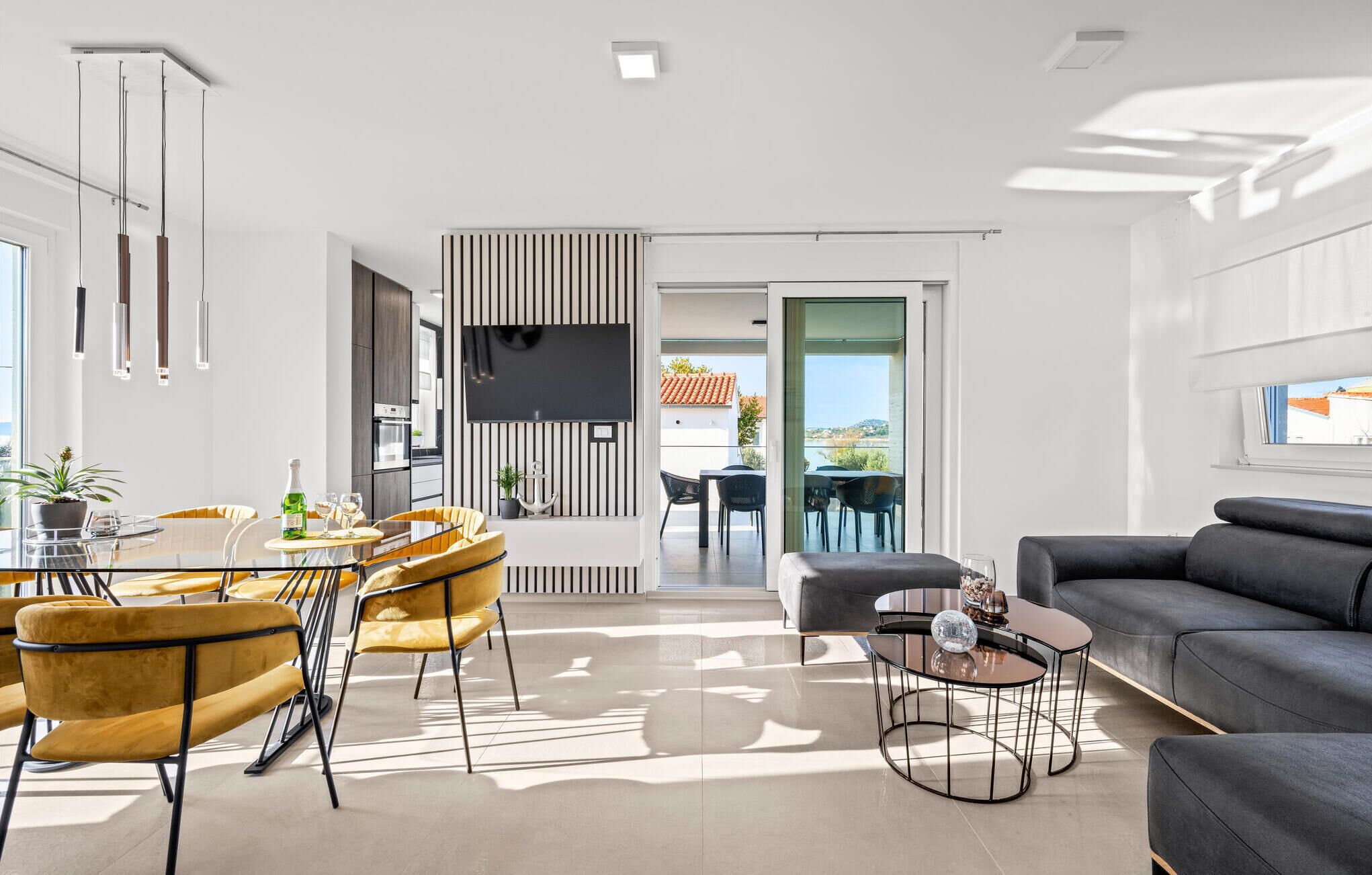 Una sala moderna con un sofá, un comedor con sillas y una mesa, un televisor montado en la pared, y un balcón con vistas.
