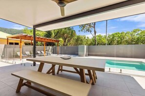 A modern outdoor dining area with a pool, clear glass walls, and a view of greenery.