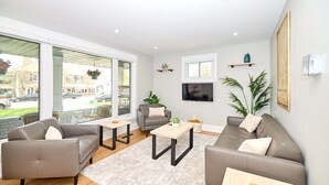 Un salon moderne avec deux canapés, une table basse et un téléviseur. De grandes fenêtres offrent une vue sur la rue.