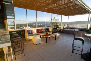 Amplia zona de estar al aire libre con vistas a la ciudad, una pérgola de madera y una zona de bar con taburetes.