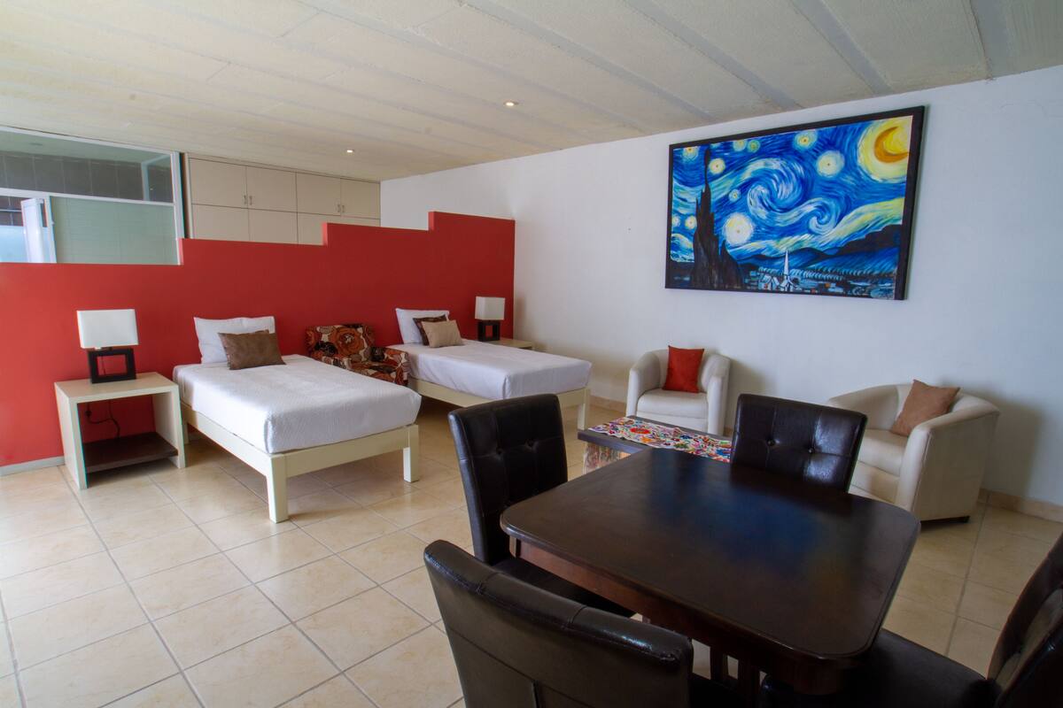 A hotel room with a bed, two bedside tables, a chair, a sofa, a dining table, and a large painting on the wall.
