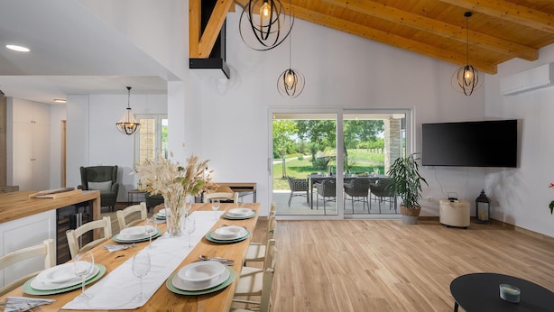 Un espace repas moderne avec une table en bois disposée pour six personnes, un téléviseur fixé au mur et une vue sur l’extérieur grâce à de grandes portes coulissantes.