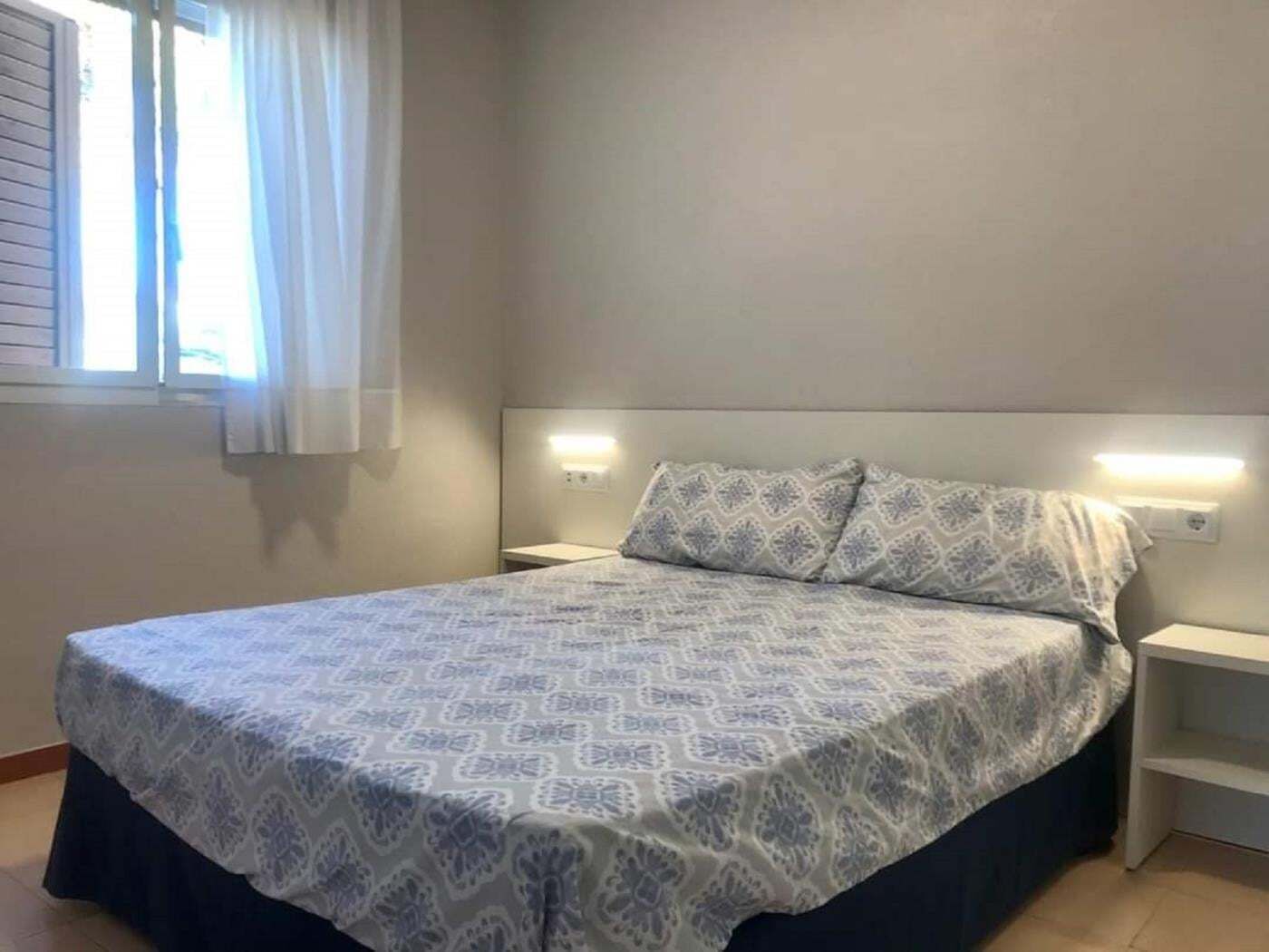 Een enkel bed met een gebedruid sprei, twee nachtkastjes met lampen, een raam met gordijnen, en een effen muur.