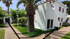 Un bâtiment blanc, avec un palmier, une entrée fermée par un portail et un chemin piétonnier dallé.