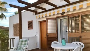 Un balcon avec une table et des chaises blanches, un fauteuil rembourré et une porte en bois avec des volets.