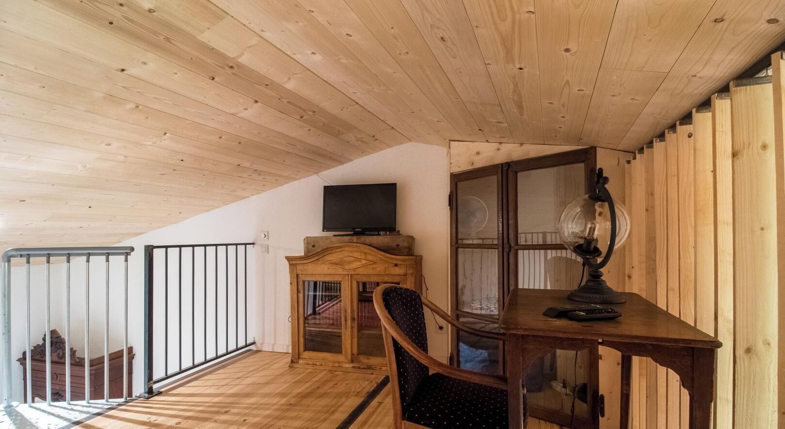 Une pièce avec un plafond en bois, un téléviseur posé sur un meuble, un bureau avec une lampe et une chaise.