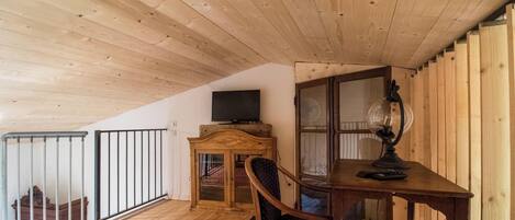 A room with a wooden ceiling, a television on a cabinet, a desk with a lamp, and a chair.