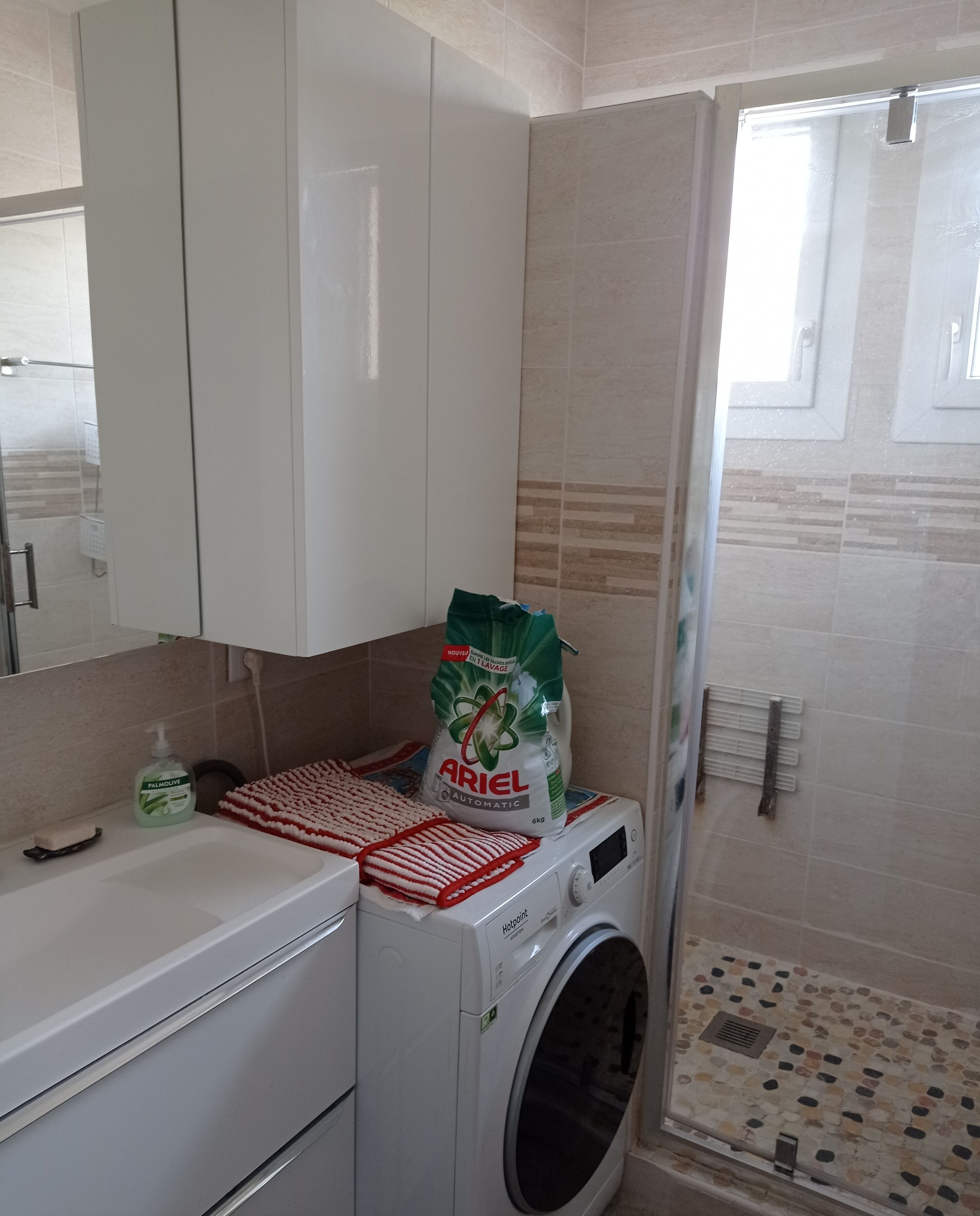 A compact laundry area with a washing machine, a drying rack, and a bag of Ariel detergent.
