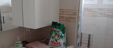 A compact laundry area with a washing machine, a drying rack, and a bag of Ariel detergent.