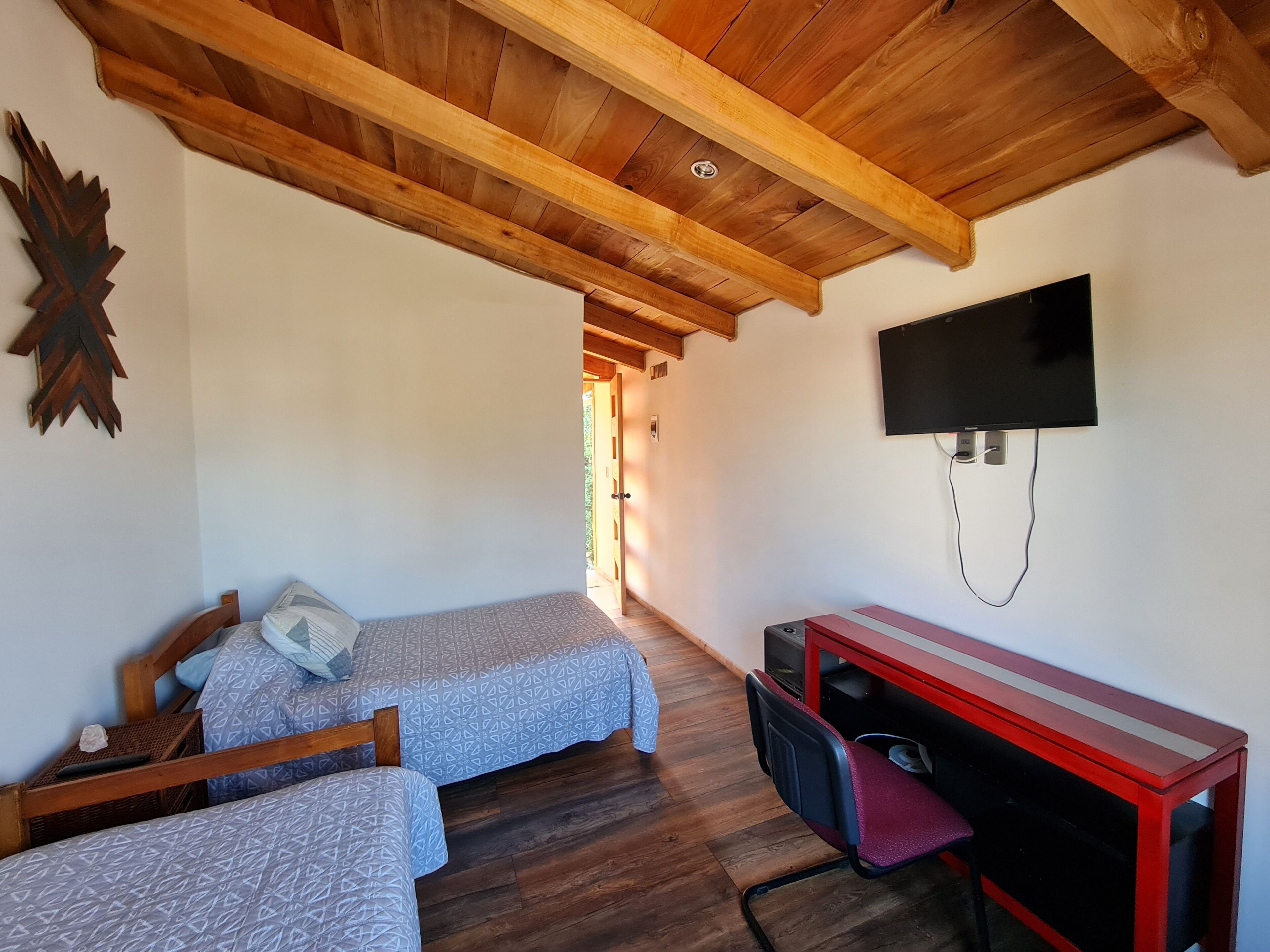 A room with two single beds, a wooden ceiling, a TV mounted on the wall, and a desk with a chair.