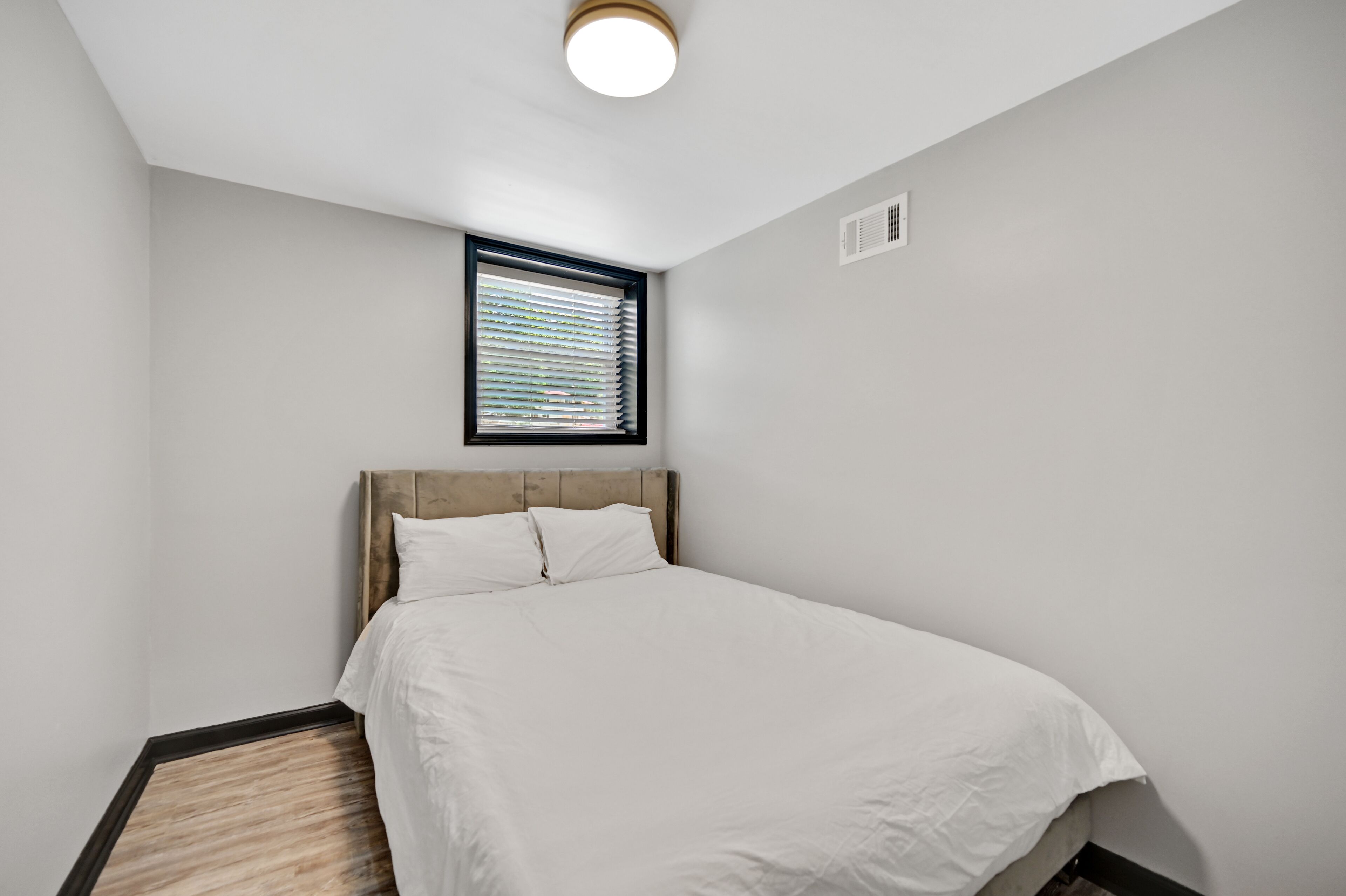 Une chambre à coucher avec un lit, une fenêtre avec des stores, un luminaire au plafond et une bouche d’aération fixée au mur.