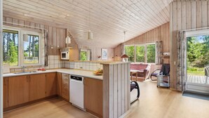 A modern kitchen with wooden cabinets, a built-in microwave, and a dishwasher. There is a dining area with a table and chairs, and large windows offering views of the outdoors.