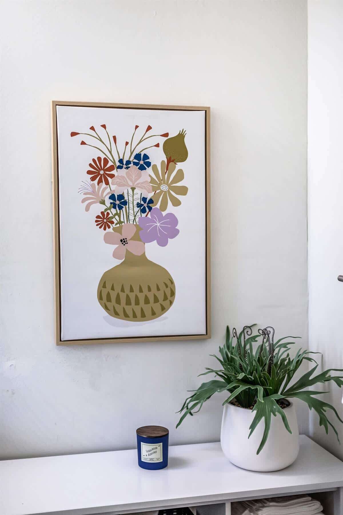 Una obra d'art floral emmarcada penja d'una paret blanca, hi ha una planta en test i una espelma sobre una taula consola blanca.