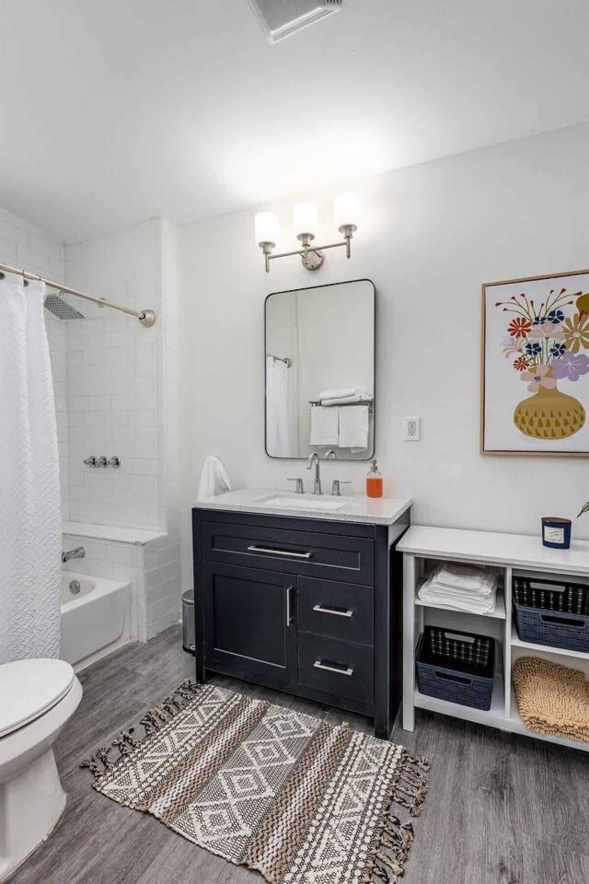 A modern bathroom with a white toilet, a black vanity with a mirror, a shower, and a patterned rug.