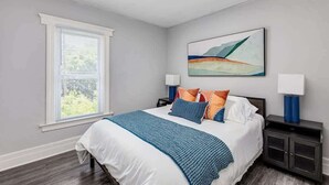 A bedroom with a bed, a window, a painting, and a nightstand. - 5BR - 12 Mins to Downtown 6 Queens + Free Parking (Detroit)