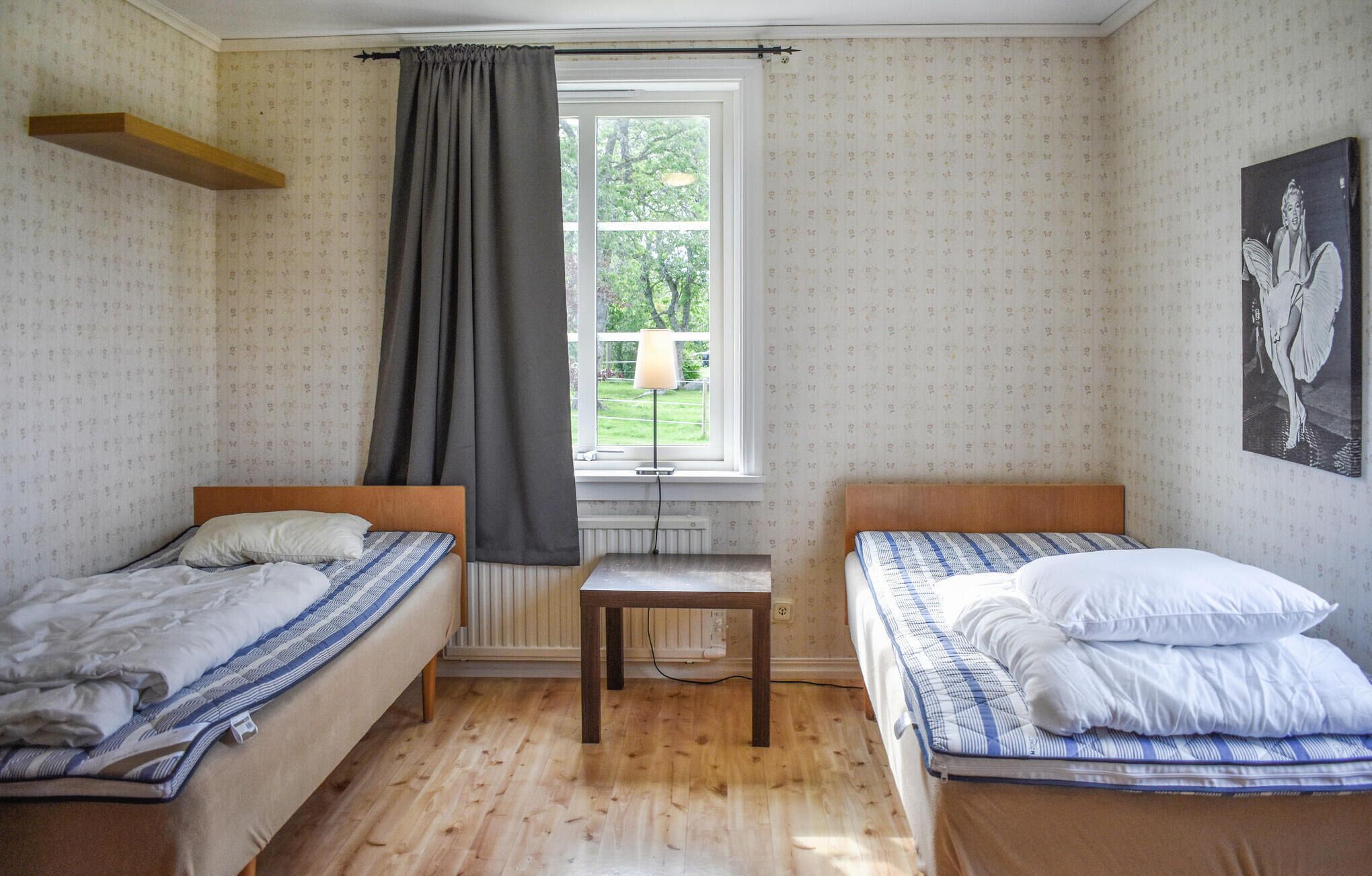 A room with two single beds, a wooden floor, a window with a view of greenery, and patterned wallpaper.