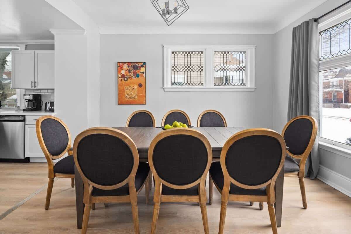 Uma sala de jantar com mesa e cadeiras de madeira, cozinha ao fundo e uma janela com vidro de grade.