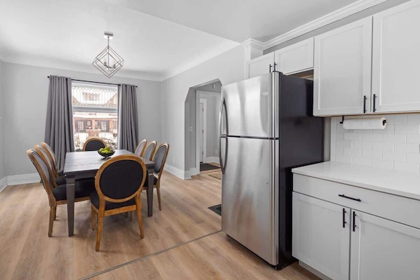 A modern kitchen with white cabinets, a stainless steel refrigerator, and a dining area with a table and chairs.