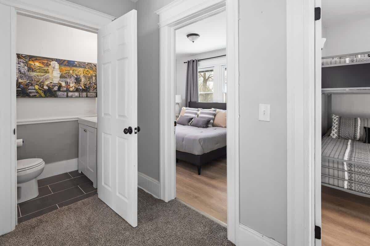 Een gang die leidt naar een slaapkamer met een bed, een badkamer met toilet, en een groot schilderij aan de muur.