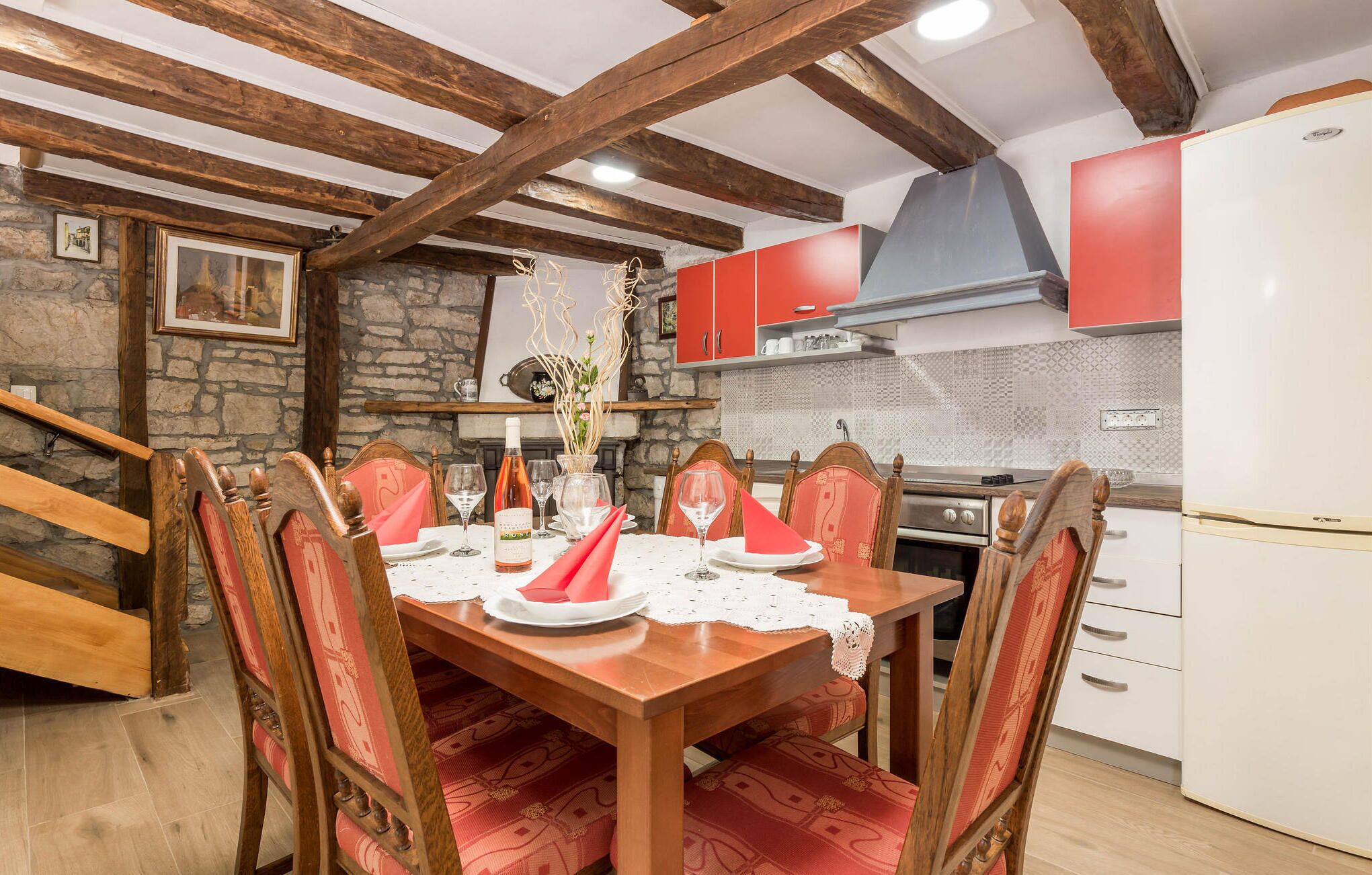Un coin repas avec une table en bois dressée pour quatre personnes, entourée de chaises dotées de coussins rouges. On aperçoit un mur en pierre et une cheminée à l’arrière-plan.