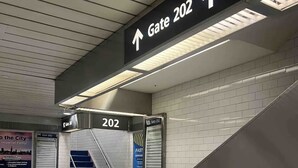 Escada rolante levando para o Portão 202 em uma estação de metrô com piso ladrilhado e sinalização.