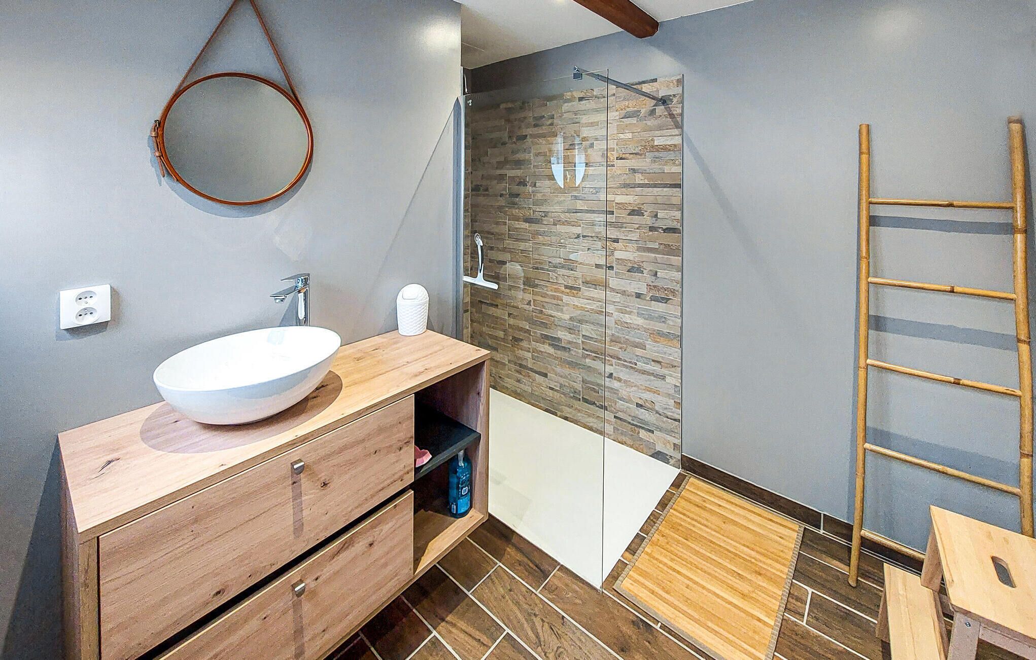 A modern bathroom with a wooden vanity, a large mirror, a stone shower wall, and a wooden ladder.