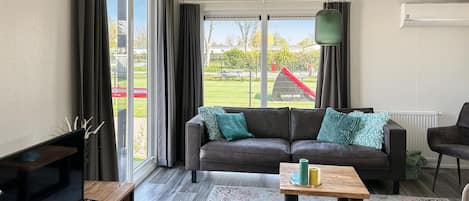 Ett modernt vardagsrum med en mörk lädersoffa, ett träbord och en tv som står på ett träställ. Stora fönster med gardiner ger utsikt över en park och en lekplats.