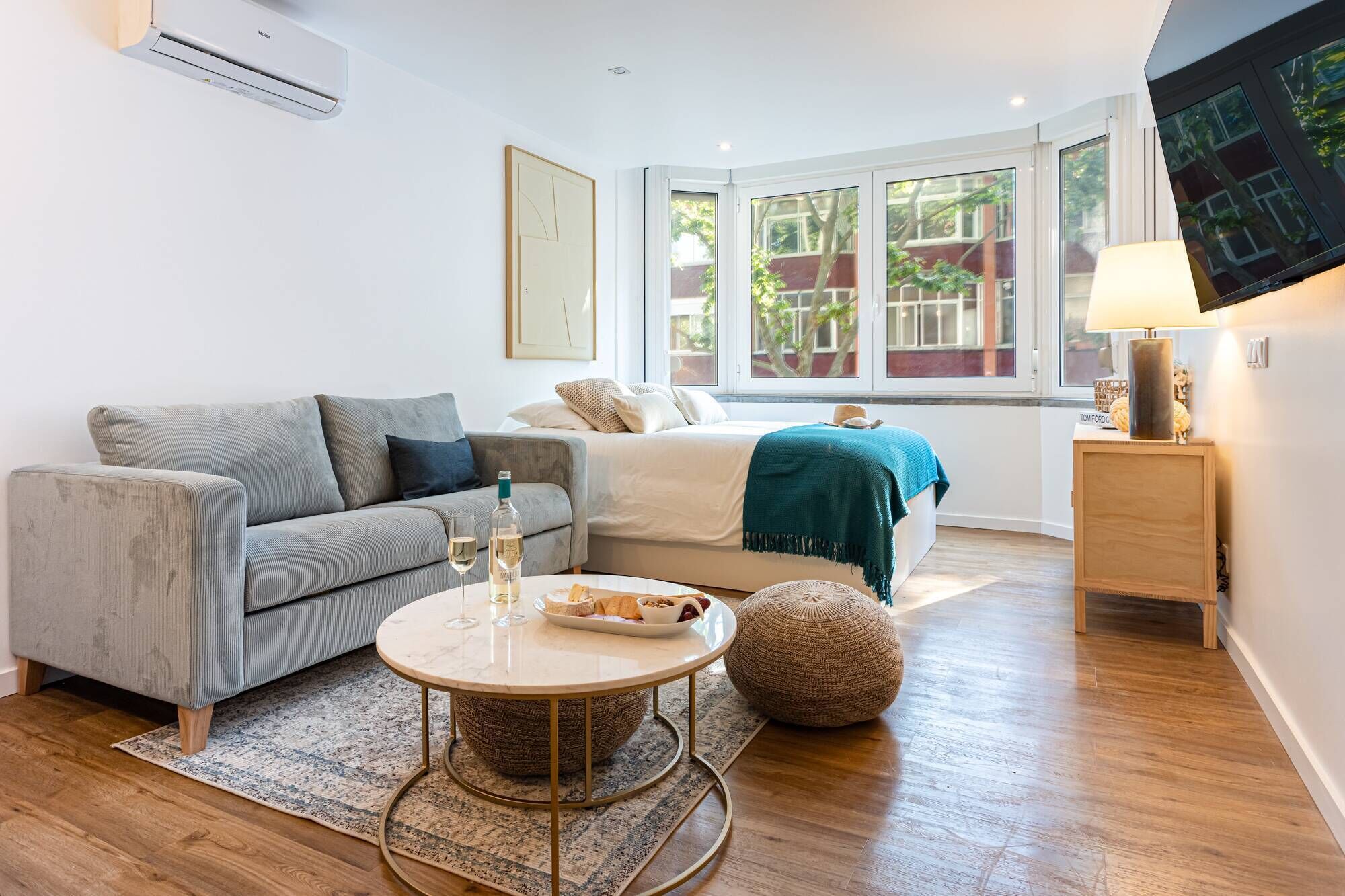 Ein modernes, gut beleuchtetes Zimmer mit einem grauen Sofa, einem Bett mit einer türkisfarbenen Decke, einem runden Couchtisch mit Flasche und Gläsern, einem gewebten Pouf und einem Fernseher an der Wand.