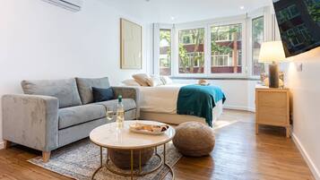 Ein modernes, gut beleuchtetes Zimmer mit einem grauen Sofa, einem Bett mit einer türkisfarbenen Decke, einem runden Couchtisch mit Flasche und Gläsern, einem gewebten Pouf und einem Fernseher an der Wand.