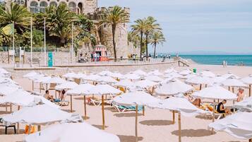 Ein Strand mit Reihen weißer Sonnenschirme und Liegestühle, ein Steinhaus mit einem Turm und im Hintergrund Palmen.