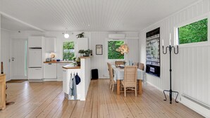 Un intérieur lumineux et blanc, avec un plancher en bois, un coin cuisine doté de meubles blancs, une table à manger entourée de chaises en osier et une grande fenêtre donnant sur un espace verdoyant.