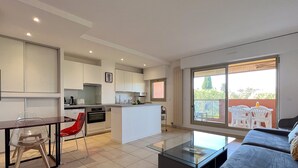 A modern apartment with a kitchen, dining area, and living room. It features a large sliding glass door to the balcony, a glass coffee table, and a sofa. - LA BADINE Studio AirCo Pool for 2 By Sunset (Antibes)