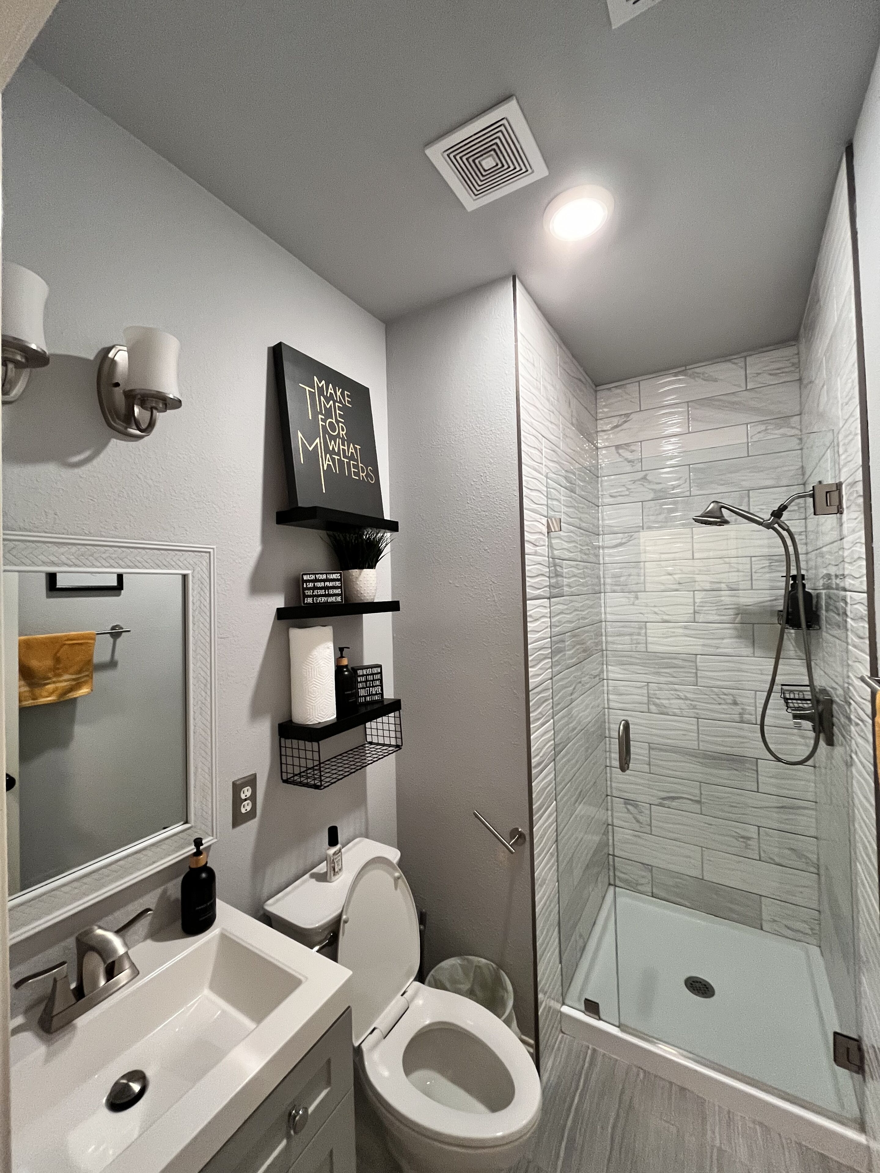 A modern bathroom with a toilet, sink, and shower. There are shelves with toiletries and a sign on the wall.