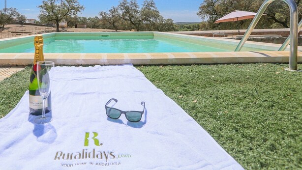 Een zwembadterras met een fles Moët & Chandon Brut champagne, een glas en een paar zonnebrillen op een handdoek met het logo "Ruraldays.com" en de tekst "Your home in Andalucía".
