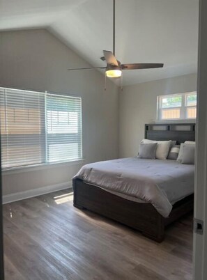 Relax in this cozy bedroom with a large bed and ceiling fan, featuring two windows with blinds.