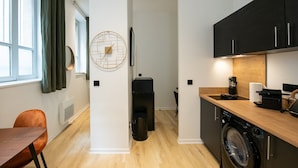 A compact kitchen with a washing machine, a coffee machine, and a dining area with a wooden table and chair. - The O1 - room 1 - Opera - 26m² (Lyon)