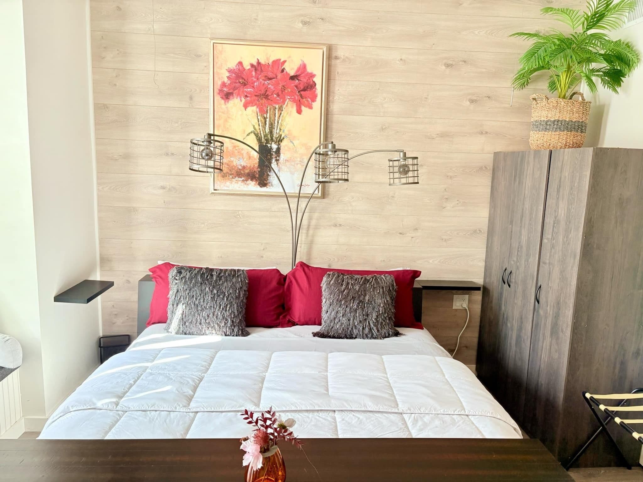 A bedroom with a bed, pillows, a bedside table, a nightstand, a wall-mounted picture, a plant, and a wardrobe.