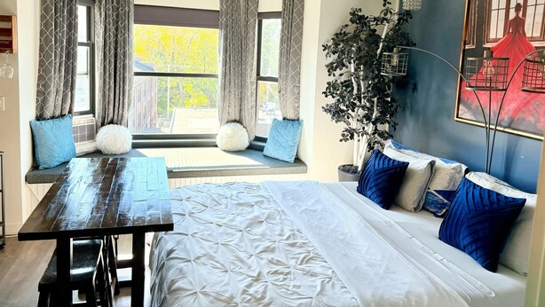 Ein modernes Schlafzimmer mit einem großen Bett, einem Esstisch und Blick auf Bäume durch die Fenster.