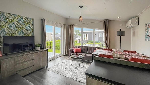 Een moderne woonkamer met een flatscreen televisie, een bank, een eettafel met rode kussens en een groot raam met uitzicht op buiten.