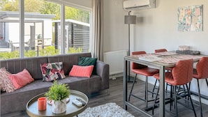 Een moderne woonkamer met een grijze bank, rode kussens, een ronde salontafel met een plant en een afstandsbediening, een eettafel met rode stoelen en een groot raam met uitzicht op gebouwen en groen.